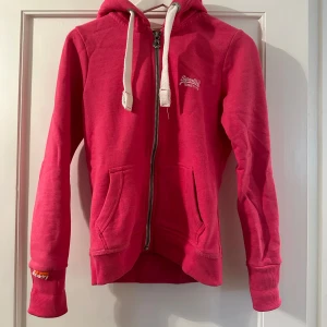 Rosa hoodie från Superdry - Säljer en rosa hoodie från Superdry med dragkedja och broderad logga på bröstet.