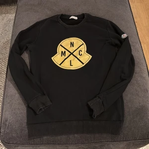 Svart tröja från Moncler - Säljer en svart tröja från Moncler med ett stort gult tryck på framsidan. Tröjan har långa ärmar och ribbade muddar vid ärmslut och nederkant. Perfekt för en stilren look.