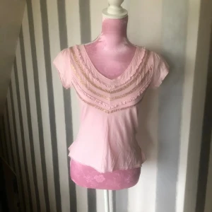 Rosa topp med volangdetaljer - Söt rosa topp med korta ärmar och v-ringning. Toppen har fina volangdetaljer och glittrande band både framtill och baktill. Perfekt för en feminin och lekfull stil.