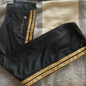 Svarta byxor med guldiga detaljer - Skit snygga lågmidjade strait legged byxor. Sitter tight men bekvämt upptill och svänger i benen. Långa ben med guld ränder längst sidan. Passar för träning och annat. 