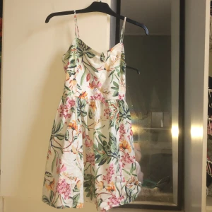 Blommig sommarklänning från H&M - Säljer en söt blommig klänning från H&M med smala axelband och ett färgglatt mönster av blommor i rosa, orange och grönt. Perfekt för varma sommardagar. Klänningen har en luftig passform och är knälång.
