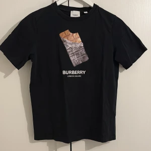 Svart t-shirt från Burberry - Säljer en svart t-shirt från Burberry med ett coolt tryck av en chokladkaka på framsidan. T-shirten är gjord i mjukt bomullsmaterial. Perfekt för en stilren och avslappnad look. Tröjan är i bra skick samt inköpt från NK kids i Stockholm.