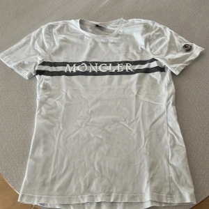 Vit t-shirt från Moncler - Säljer en vit t-shirt från Moncler med korta ärmar och ett tryck av märkesnamnet i grått över bröstet. T-shirten har en broderad logga på ärmen. Perfekt för en stilren look.