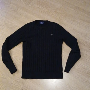 Svart stickad tröja från Gant - Säljer en stilren svart stickad tröja från Gant med ribbad struktur. Tröjan har långa ärmar och en liten broderad logotyp på bröstet. Perfekt för kyligare dagar.