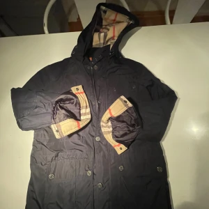 Svart parkajacka från Burberry - Säljer en stilren svart parkajacka från Burberry med klassiskt rutigt foder i beige, mörkblå är den . Jackan har en huva och knappar framtill. Perfekt för kyliga dagar med sin quiltade insida.