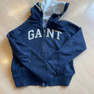Mörkblå hoodie från GANT - Säljer en snygg mörkblå hoodie från GANT med dragkedja och stor logga på bröstet. Tröjan har långa ärmar och en justerbar huva med snören. Perfekt för en avslappnad stil.