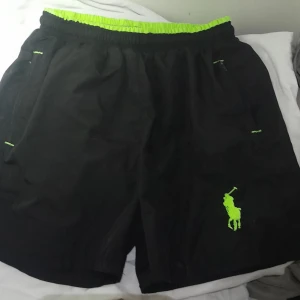 Ralph Lauren Polo shorts  - Snygga svarta shorts från Ralph Lauren Polo med neongröna detaljer och logga. De har en elastisk midja och är perfekta för en sportig look. Shortsen är tillverkade i ett lätt och bekvämt material.