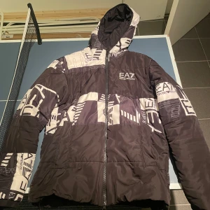 Svart och vit pufferjacka från Emporio Armani - Säljer en svart och vit pufferjacka från Emporio Armani med ett unikt mönster och logotypdetaljer. Jackan har en dragkedja framtill och en huva för extra skydd. Perfekt för kyligare dagar. Jackan är i bra skick och inte trasig någonstans.