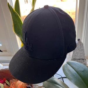 Svart keps från New Era - Säljer en svart keps från New Era med LA-logga framtill. Kepsen är i modellen 9FIFTY och är tillverkad av bomull. Perfekt för en stilren look.