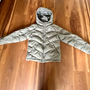 Grå dunjacka från Nike - Säljer en snygg grå dunjacka från Nike. Jackan har en huva och dragkedja framtill. Perfekt för kyligare dagar med sin vadderade design. Den har en klassisk look med Nike-loggan på bröstet. Bra skick, säljer då den blivit för liten😊