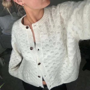 Vit stickad kofta med knappar - Mysig vit stickad kofta med ett diskret mönster och bruna knappar. Perfekt för kyliga dagar och ger en stilren look. Passar bra till både jeans och kjol.