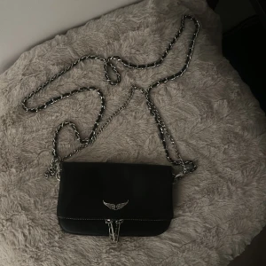 Svart axelväska med kedja - Snygg svart axelväska från Zadig & Voltaire med silverfärgad kedja och dragkedja. Väskan har en stilren design med en liten logotypdetalj framtill. Perfekt för att bära det nödvändigaste med stil. Väska har inga defekter alltså är den i nyskick, man kan ta bort axelbandet om man föredrar att ha som en handväska. Det är den minsta modellen. Hör av dig vid funderingar och pris kan alltid diskuteras