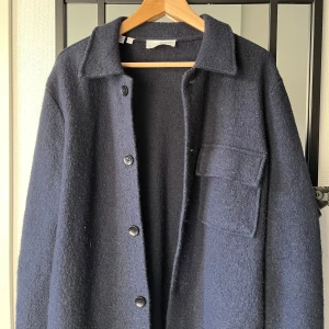 Mörkblå stickad overshirt - Snygg mörkblå overshirt i 100% ull med knappar framtill och en bröstficka. Den är i storlek S men lite lång i armarna. Köpt för 1200 kr från another store, märke selected homme. Inga slitage.