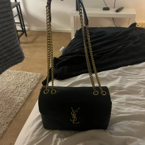 Svart quiltad axelväska med kedja - Snygg svart quiltad axelväska från YSL med guldfärgad kedja och logotyp. Väskan har en elegant design och är perfekt för att ge din outfit en lyxig touch. Den är tillverkad i skinn och har en stilren look som passar till många tillfällen.