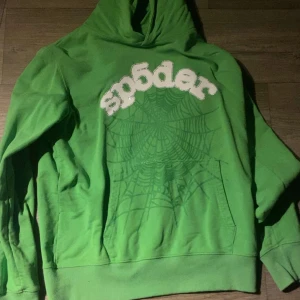 Grön Sp5der Hoodie - Säljer en grön hoodie med en cool spindelnätsdesign och texten 'spöder' på framsidan. Perfekt för en avslappnad stil. Den har en klassisk känguruficka och en bekväm huva.