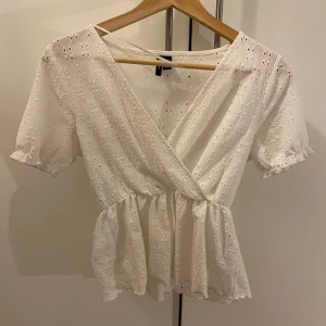 Vit omlottblus med spetsdetaljer - Säljer en söt vit omlottblus med spetsdetaljer och korta ärmar. Blusen har en volangkant nedtill och ett fint broderat mönster. Perfekt för en somrig look!Knappt använd💕💕