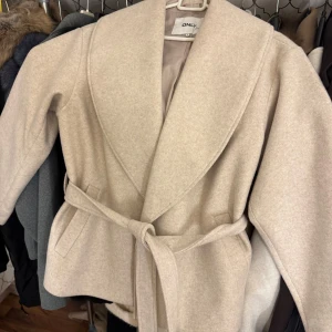 Beige kappa från ONLY - Snygg beige kappa från ONLY med ett elegant bälte i midjan. Den har en stilren design med långa ärmar och bred krage. Perfekt för höst och vår!
