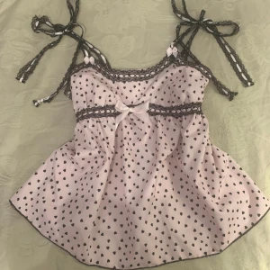 Super söt ljus rosa topp  - Ljus rosa topp med svarta hjärtan och spetsdetaljer. Den har rosetter på axelbanden och en söt rosett framtill, den sitter fast med en säkerhetsnål vilket inte syns så mycket, men den är enkel att sy fast om man vill! Bra skick💕💕💕