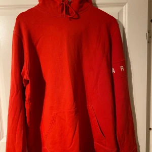 Röd hoodie från Axel Arigato - Säljer en snygg röd hoodie från Axel Arigato. Den har en justerbar huva med snören och en stor ficka framtill. På ärmen finns textdetaljer. Perfekt för en avslappnad stil.
