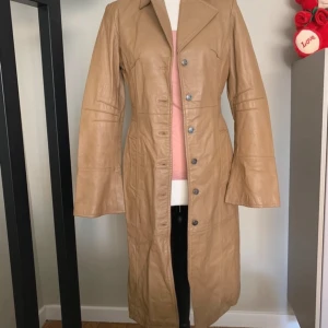 Beige skinnkappa S  - Snygg beige skinnkappa med knappar framtill och klassisk krage. Långärmad design med en stilren look som passar perfekt för höst och vår. Kappan har en figursydd passform och knappar även vid ärmsluten. Fin camel färg 