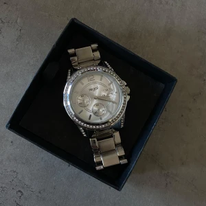 Silverfärgad klocka från Regal - Elegant silverfärgad klocka från Regal med glittriga detaljer runt urtavlan. Klockan har en metallarmband och tre små urtavlor inuti den större urtavlan.  Oanvänd &  mycket bra skick.