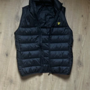 Svart dunväst från Lyle & Scott - Snygg svart dunväst från Lyle & Scott med dragkedja och ett broderat gult emblem på bröstet. Funkar perfekt nu på våren och sommaren! Passformen säger xs men skulle säga att den passar både s och m. Köpt för 1200 säljer nu för ett bra pris på 550!