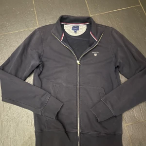 Mörkblå tröja med dragkedja från Gant - Säljer en stilren mörkblå tröja från Gant med dragkedja och broderad logga på bröstet. Tröjan har långa ärmar och ribbade muddar vid ärmslut och nederkant. Perfekt för en avslappnad look.