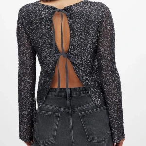 Svart glittrig topp med öppen rygg - Snygg svart glittrig topp med långa ärmar och en fantastisk öppen rygg med knytband. Perfekt för att sticka ut på festen. Den har en elegant och modern design som fångar ljuset på ett vackert sätt. Med lappen på 