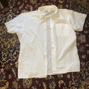 Vit kortärmad skjorta från John Noble - Säljer en klassisk vit kortärmad skjorta från John Noble. Skjortan har en bröstficka och knappar framtill. Perfekt för en stilren look.