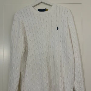 Vit stickad tröja från Polo Ralph Lauren - Säljer en stilren vit stickad tröja från Polo Ralph Lauren med kabelstickat mönster och den klassiska loggan broderad på bröstet. Tröjan har långa ärmar och ribbade muddar vid ärmslut och nederkant. Perfekt för en elegant och avslappnad look.
