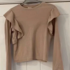 Beige tröja med volangdetaljer - Säljer en stilren beige tröja med volangdetaljer på ärmarna. Tröjan har långa ärmar och en rund halsringning, perfekt för en chic och avslappnad look.