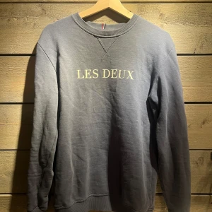 Grå sweatshirt från Les Deux - Säljer en stilren grå sweatshirt från Les Deux med tryck på bröstet. Tröjan har långa ärmar och ribbade muddar vid hals, ärmslut och nederkant. Perfekt för en avslappnad look.