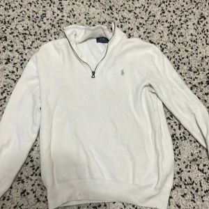 Vit tröja med dragkedja från Polo Ralph Lauren - Säljer en stilren vit tröja från Polo Ralph Lauren med dragkedja vid halsen. Tröjan har långa ärmar och en liten broderad logga på bröstet. Perfekt för en klassisk och avslappnad look.