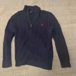 Mörkblå stickad tröja från Ralph Lauren - Säljer en snygg mörkblå stickad tröja från Ralph Lauren med dragkedja vid halsen och det klassiska röda logotypbroderiet på bröstet. Tröjan har långa ärmar och ribbade muddar för en stilren look. Perfekt för kyligare dagar!
