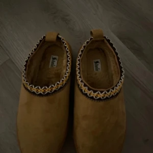 Bruna mockaskor med mönstrad kant - Säljer ett par bruna mockaskor med en snygg mönstrad kant i svart och gult. Skorna har en rund tå och en bekväm platt sula. Perfekta för en avslappnad stil.