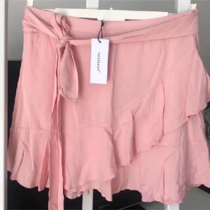 Rosa kjol från Vovelles M - Söt rosa kjol från Vovelles med volangdetaljer och knytband i midjan. Perfekt för en somrig look. Kjolen har en omlott-design som ger en fin passform. Midjemått 78 cm 