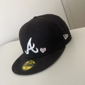 Svart keps från New Era - Snygg svart keps från New Era med broderad vit logotyp och ett litet hjärta med baseballmotiv. På sidan finns texten 'Love & Hustle' i vitt och rött. Kepsen har en platt skärm och är perfekt för sportiga tillfällen.