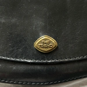 Svart axelväska från The Trend - Säljer en stilren svart axelväska från The Trend. Väskan är tillverkad i skinn och har en smal axelrem. Insidan är fodrad med ett mönstrat tyg och har en logotypetikett. Perfekt för att bära med sig det nödvändigaste.