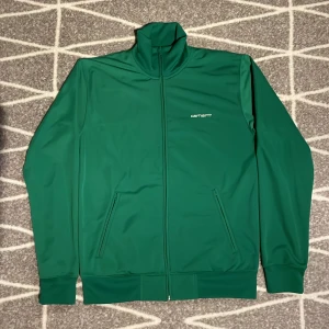 Grön jacka från Carhartt - Säljer en snygg grön jacka från Carhartt i storlek M. Jackan har en dragkedja framtill och är tillverkad i 100% polyester. Perfekt för en stilren look med långärmade ärmar och en enkel design.