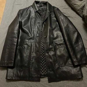 Svart skinnjacka från Urban Outfitters - Snygg svart skinnjacka från Urban Outfitters med quiltad insida och knappar framtill. Jackan har en klassisk design med långa ärmar och stora fickor. Perfekt för en stilren look.