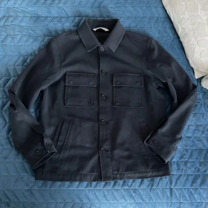 Mörkblå overshirt från Zara - Säljer en stilren mörkblå overshirt från Zara med klassisk krage, två stora bröstfickor med lock och knäppning framtill. Jackan har långa ärmar med knapp vid ärmslut och raka linjer för en clean look. Perfekt att slänga över en hoodie eller t-shirt.