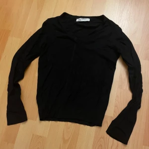 Svart v-ringad tröja från Zara - Svart långärmad tröja från Zara med v-ringning och slits vid ärmsluten. Tröjan är i mjukt material och har en klassisk, enkel look som funkar till många outfits. Perfekt för dig som gillar stilrena basplagg med en liten twist.