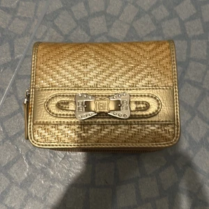 Guldig plånbok med metallrosett - Snygg guldig plånbok från L.Credi med präglat mönster och en cool metallrosett på framsidan. Plånboken har flera kortfack, fack för ID och myntfack med dragkedja. Perfekt för dig som vill ha något som sticker ut i väskan.