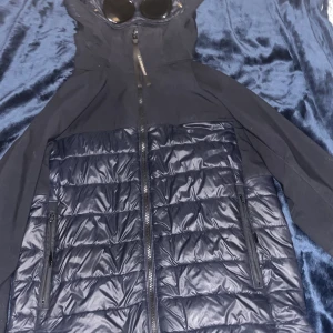 Svart pufferjacka från C.P company - Cool svart pufferjacka med quiltad front och släta ärmar. Jackan har hel dragkedja, två sidofickor med dragkedja och en huva med inbyggda glasögon för extra edge. Perfekt för dig som vill sticka ut i vinterkylan.