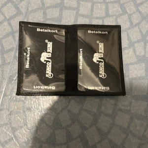 Svart plånbok från Banco Rhino - Klassisk svart plånbok i skinn från Banco Rhino. Plånboken har flera fack för kort och sedlar samt ett transparent fönster för ID eller foto. Stilren design med präglad logga på framsidan och smidig storlek som passar i fickan.