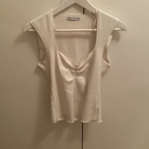 Vit ärmlös topp från Zara - Snygg vit topp från Zara med breda axelband och djup v-ringning. Toppen har en lätt rynkad detalj framtill som ger en cool twist. Perfekt till jeans eller kjol för en fräsch look. Materialet känns mjukt och följsamt.