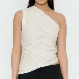 Vit oneshoulder topp i spets - Stilren vit topp med oneshoulder-design och lätt asymmetrisk nederkant. Tunt spetsmaterial med diskret blommönster ger en romantisk känsla. Perfekt för dig som vill sticka ut med en unik och trendig look.