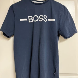 Marinblå BOSS t-shirt i bomull - Snygg marinblå t-shirt från BOSS med vit logga framtill. Klassisk rund halsringning och korta ärmar. Tillverkad i mjuk bomull som känns skön mot huden. Perfekt för dig som gillar stilrena och trendiga plagg med premiumkänsla.