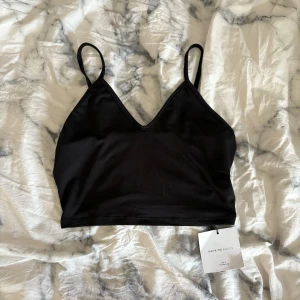 Svart linnetopp från SHEIN Basics  - En svart, enkel och stilren linnetopp från SHEIN Basics i storlek XS. Toppen har smala axelband och v-ringad hals, perfekt för lager-på-lager eller som den är. Gjord i mjukt och stretchigt syntetmaterial för en bekväm passform.