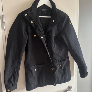 Svart jacka från Peak Performance  - Svart jacka från Peak Performance med guldiga knappar och dragkedja. Jackan har flera fickor framtill, hög krage och klassisk passform. Perfekt för dig som vill ha en stilren och funktionell look till höst och vår.